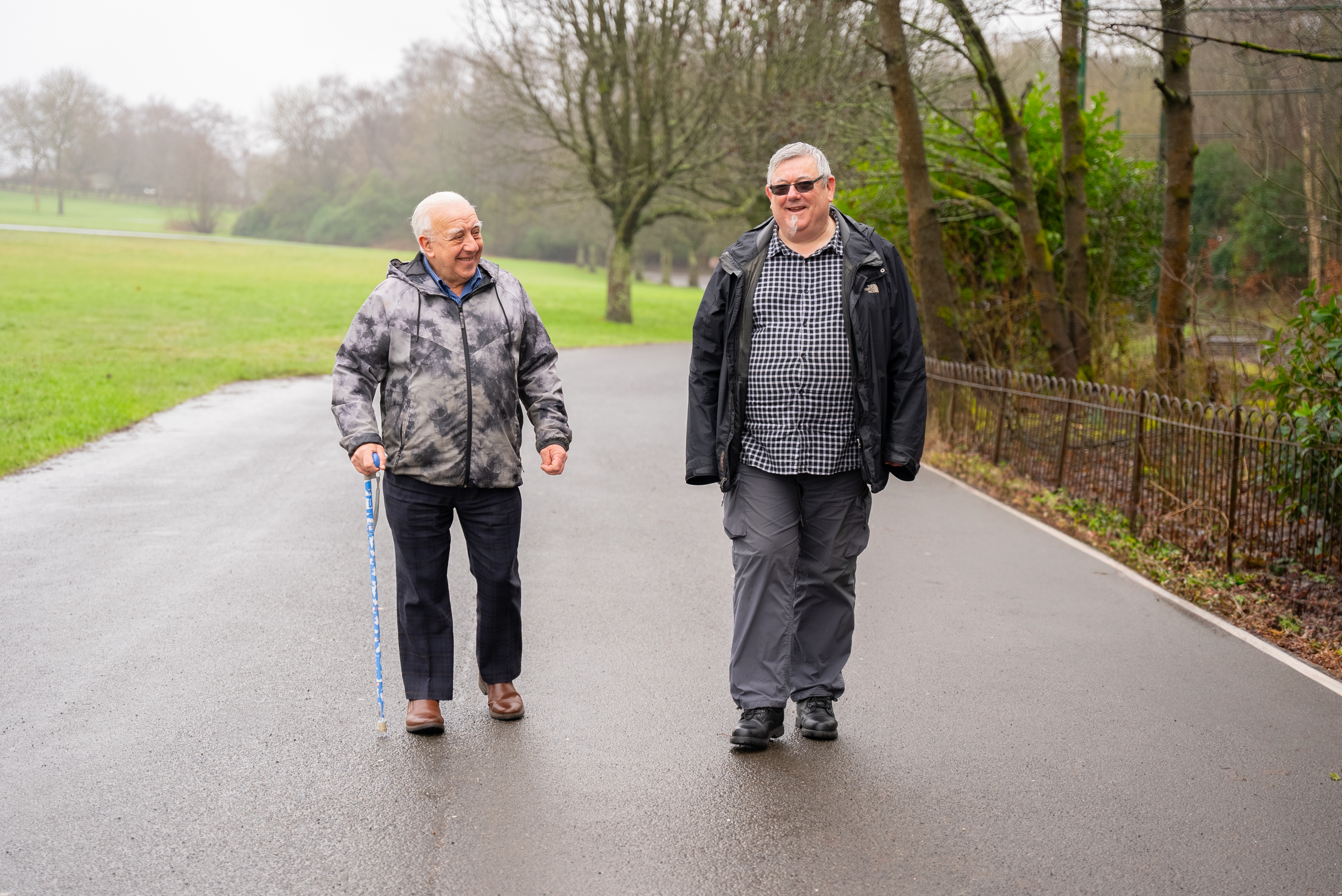 Two older men walking together in the park. One has a walking stick. Both are smiling