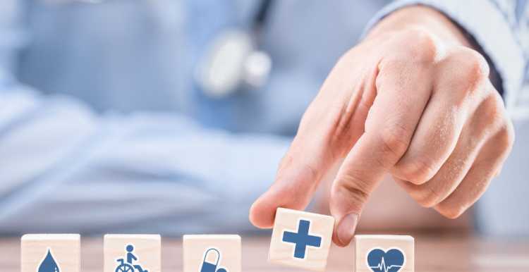A doctor with stethoscope with mini blocks in front of him representing health care and his hand picking up one with a blue cross on it 