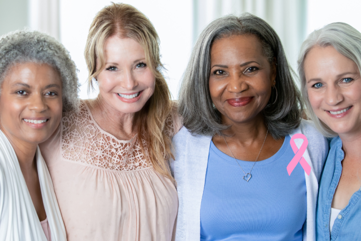 A photo of four middle aged women smiling at the camera with a pink ribbon motif in the bottom right corner