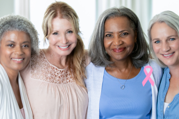 A photo of four middle aged women smiling at the camera with a pink ribbon motif in the bottom right corner