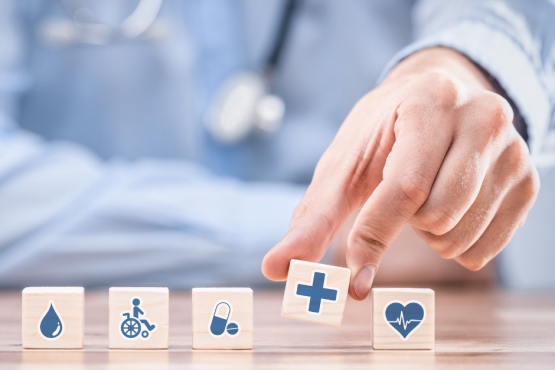 A doctor with stethoscope with mini blocks in front of him representing health care and his hand picking up one with a blue cross on it 
