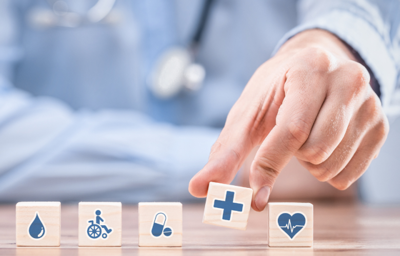 A doctor with stethoscope with mini blocks in front of him representing health care and his hand picking up one with a blue cross on it 